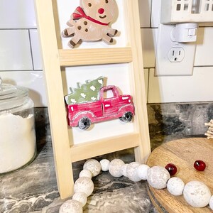 May include: A wooden ladder shelf with two tiers, featuring a red truck carrying a green Christmas tree and a reindeer with a red bow. The ladder shelf is decorated with a white bead garland and red berries.
