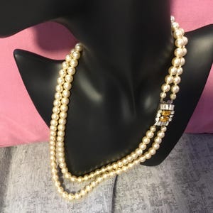 Op de afbeelding: Een dubbele rij parelketting met een gouden en heldere strass sluiting. De ketting is op een zwart manikin.