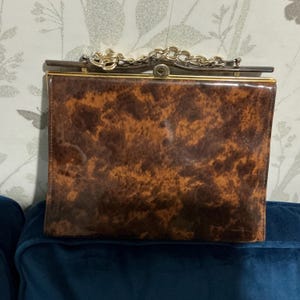 Op de afbeelding: Een vintage bruine handtas met een schildpadpatroon. De rechthoekige tas heeft een goudkleurig frame en een kettingriem. De sluiting is goudkleurig. De tas ligt op een blauw oppervlak.