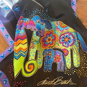May include: A black scarf featuring a colorful graphic print of three dogs decorated with floral patterns, polka dots, and chevron stripes in shades of blue, green, pink, and orange. The signature Laurel Burch is printed in gold at the bottom.