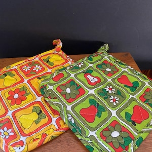May include: Two square quilted pot holders featuring a repeating grid pattern of flowers and pears. One pot holder displays yellow pears and orange flowers on a yellow background, while the other shows red pears and green flowers on a green background.
