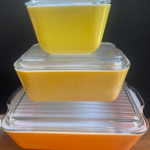 May include: A stack of three vintage Pyrex dishes. The top dish is yellow, the middle dish is a darker yellow, and the bottom dish is orange. Each dish has a clear glass lid. The dishes are rectangular and have a retro aesthetic.