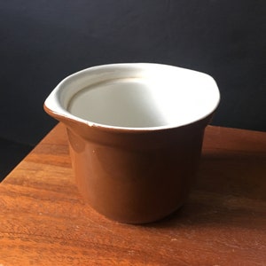 May include: A brown ceramic creamer with a white interior. The creamer is sitting on a wooden surface.