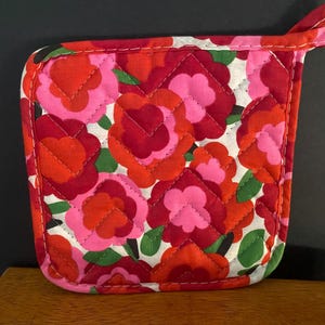 May include: A square quilted pot holder featuring a vibrant floral pattern with overlapping red, orange, and pink flower shapes on a white background, accented by small green leaves. The fabric is stitched in a diamond pattern with a fabric loop.