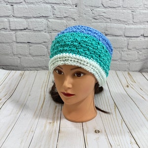 Könnte beinhalten: Eine gehäkelte Mütze mit einem weißen, türkisfarbenen und hellblauen Streifenmuster. Die Mütze befindet sich auf einem Mannequin-Kopf.