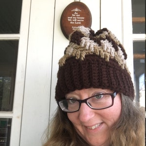 Pode incluir: Uma mulher usando um chapéu de crochê marrom, bege e bege claro com uma borda marrom. O chapéu tem um padrão texturizado. A mulher está sorrindo e olhando para a câmera. O fundo é uma porta branca com uma placa de madeira que diz "As for me and my house we will serve the Lord."