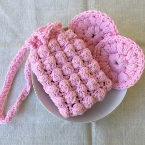 May include: Pink crocheted pouf with a textured surface and a long handle. The pouf is made of small, round stitches and has a delicate, feminine look.
