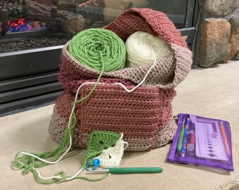 Puede incluir: Una bolsa de ganchillo de color rosa, marrón y blanco. La bolsa está llena de dos ovillos de lana, uno verde y otro blanco. También se ve un gancho de ganchillo y un pequeño cuadrado de tejido de ganchillo.