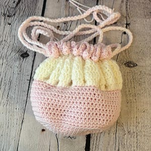 May include: A handmade crochet bag in soft pastel colors. The bag features a pink base, a cream-colored top section, and a gathered, ruffled design at the top. A matching pink crochet cord serves as a drawstring and shoulder strap.