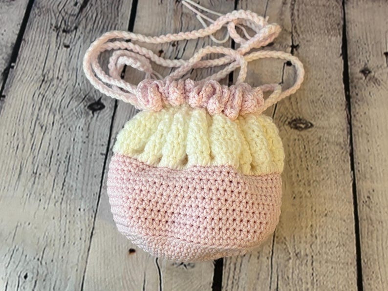 May include: A handmade crochet bag in soft pastel colors. The bag features a pink base, a cream-colored top section, and a gathered, ruffled design at the top. A matching pink crochet cord serves as a drawstring and shoulder strap.