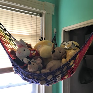 Può includere: Un'amaca a righe arcobaleno piena di vari peluche, tra cui un coniglio bianco, un'anatra gialla e un cane marrone. L'amaca è sospesa vicino a una finestra e a una porta, creando una soluzione di stoccaggio per i giocattoli di peluche.