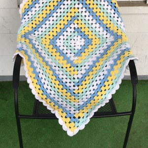 Puede incluir: Una manta de ganchillo hecha a mano con un patrón de cuadrados de la abuela. La manta presenta una combinación de colores blanco, amarillo, azul claro y verde menta. Los bordes están acabados con un borde blanco festoneado. La manta está sobre una silla de metal negro.