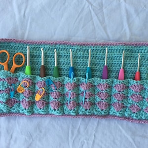 May include: A crocheted teal and purple organizer with eight slots for crochet hooks and scissors. The organizer has a scalloped design and is filled with crochet hooks and safety pins.