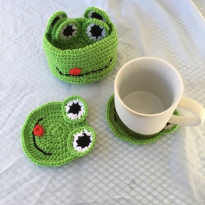 May include: Green crocheted frog-shaped coasters and a matching frog-shaped container. The coasters have black eyes with white pupils and a red mouth. The container has a similar design and is perfect for holding small items.