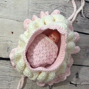 May include: A handmade crochet baby doll carrier in soft pastel colors. The carrier is primarily white with pink accents and a ruffled edge. A small plastic doll is nestled inside a pink crocheted blanket.