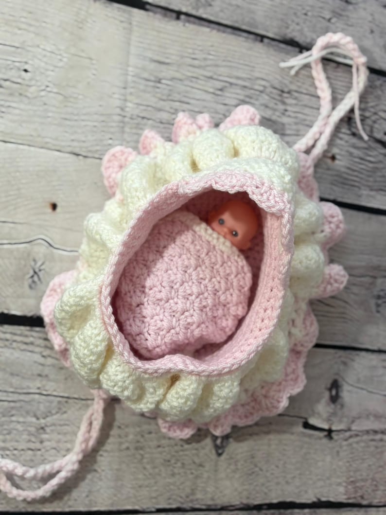 May include: A handmade crochet baby doll carrier in soft pastel colors. The carrier is primarily white with pink accents and a ruffled edge. A small plastic doll is nestled inside a pink crocheted blanket.