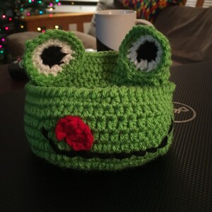 May include: Green crocheted frog hat with black eyes, white accents, and a red crocheted tongue.