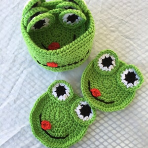 May include: Set of green crocheted frog-shaped coasters with white eyes and black pupils. The coasters are stacked inside a matching green crocheted frog-shaped container.
