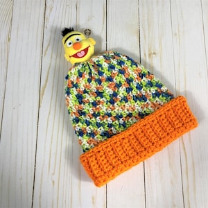 Op de afbeelding: Een gebreide muts met een kleurrijk, veelkleurig patroon. De muts heeft een feloranje rand en een geel viltpersonage met een zwarte haardos bovenop.