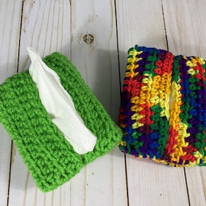 Puede incluir: Dos fundas de caja de pañuelos hechas a mano con ganchillo. Una es de color verde brillante y la otra multicolor con rojo, amarillo, azul y verde. Los pañuelos blancos son visibles en las aberturas. Las fundas son rectangulares y están diseñadas para cajas de pañuelos estándar.