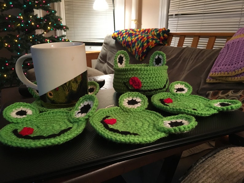 May include: Five green crocheted frog-shaped coasters with black eyes, white accents, and red mouths. A white mug with a black and green design sits on top of one of the coasters.