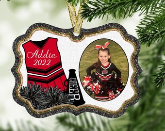 May include: A white, glitter-framed ornament with a red and black cheerleader uniform, a black megaphone with the word "CHEER" on it, and a photo of a young girl in a cheerleader uniform. The ornament is personalized with the name "Addie" and the year "2022".