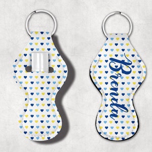 May include: Two white keychains with blue and yellow hearts. One keychain has the name "Brenda" written in blue. The other keychain has a slot for a lip balm.