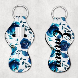 May include: A white and blue floral patterned key chain with a clear plastic pocket for lip balm or a small tube of hand sanitizer. The keychain has a black ring and the name "Emma" is printed in black on the front.