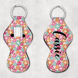May include: A pink and white floral patterned key chain with a black zippered pocket for lip balm. The keychain has a black metal ring and the name "Cassie" is printed on the front.