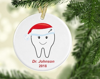 May include: White ceramic Christmas ornament with a cartoon tooth wearing a Santa hat and a toothy smile. The ornament is personalized with "Dr. Johnson 2018".