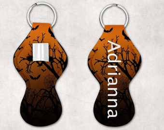 May include: Orange and black Halloween-themed key chain with a spooky forest design and the name "Adrianna" printed on it. The key chain has a loop for attaching keys and a pocket for holding a small item, such as lip balm.