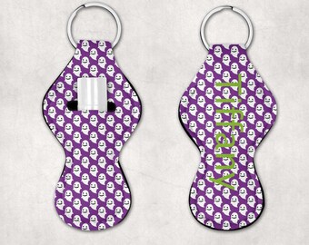 May include: Purple and white ghost-patterned lip balm holder with a silver key ring. The holder has a slot for a lip balm tube and the word "Ashley" is written in green on the front.