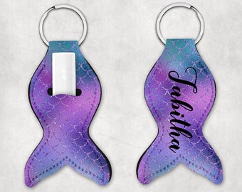 May include: A mermaid tail-shaped lip balm holder in shades of purple, blue, and teal, with a silver key ring. The holder features a scale pattern and the name "Tabitha" in black script on the back. A white lip balm is inserted in the front.