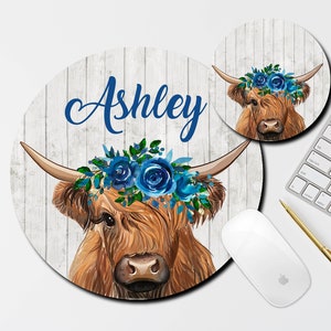 May include: A round mousepad with a white woodgrain background featuring a brown and white Highland cow with blue flowers in its hair. The cow's name, "Ashley", is written in blue text on the mousepad.