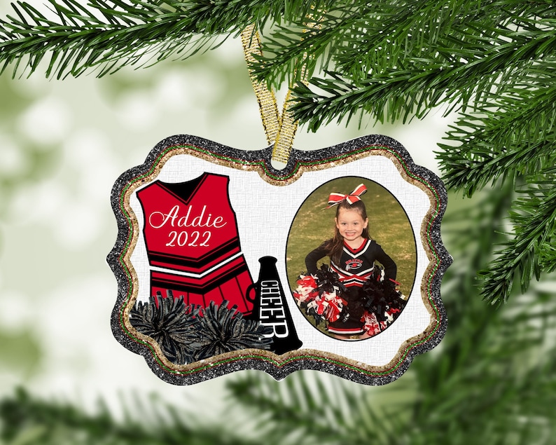 May include: A white, glitter-framed ornament with a red and black cheerleader uniform, a black megaphone with the word "CHEER" on it, and a photo of a young girl in a cheerleader uniform. The ornament is personalized with the name "Addie" and the year "2022".