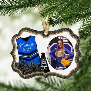 May include: A white and blue cheerleader uniform with a black megaphone and pom-poms. The uniform has the name "Mandy 2022" on the front. A photo of a cheerleader in a blue and yellow uniform is in a circle on the right side of the ornament.