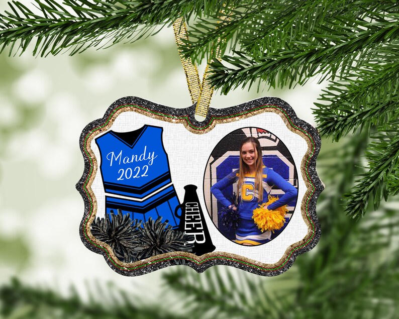 May include: A white and blue cheerleader uniform with a black megaphone and pom-poms. The uniform has the name "Mandy 2022" on the front. A photo of a cheerleader in a blue and yellow uniform is in a circle on the right side of the ornament.