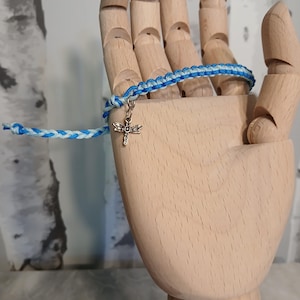 May include: A blue and white braided bracelet with a silver dragonfly charm. The bracelet is displayed on a wooden hand, showcasing the intricate knotting and the delicate charm. The bracelet is a handmade accessory.