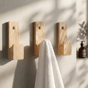 Puede incluir: Tres ganchos de pared de madera clara con un diseño rectangular vertical y una base sobresaliente. Cada gancho está fijado con un tornillo de color latón. Una toalla blanca texturizada cuelga del gancho central, mostrando su tejido suave.