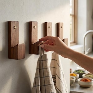 Könnte beinhalten: Vier rechteckige Wandhaken aus dunklem Holz, die in einer horizontalen Reihe montiert sind. Jeder Haken hat oben eine Messingschraube und eine geschwungene Vorderkante. Zwei Küchentücher aus Leinen, eines mit schwarzem Gittermuster und eines gestreift, hängen daran.