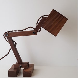 K&ouml;nnte beinhalten: Eine Schreibtischlampe aus Holz mit brauner Oberfl&auml;che. Die Lampe hat einen quadratischen Schirm und einen flexiblen Arm, der in verschiedene Positionen eingestellt werden kann.