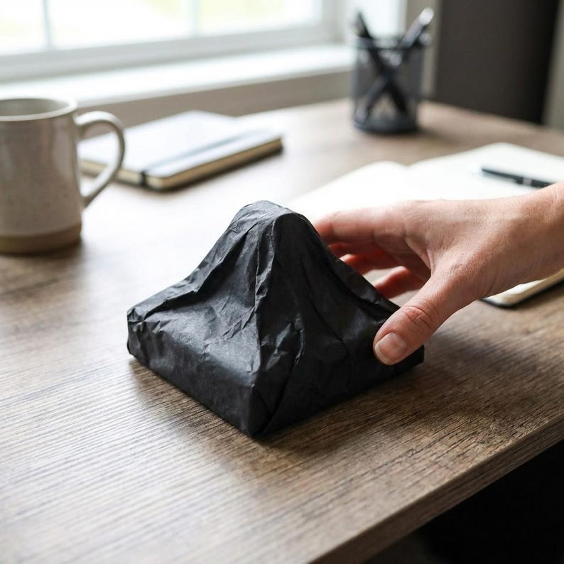 以下が含まれることがあります： 黒いピラミッド型のパッケージが、濃い色の薄紙で包まれ、木製の机の上に置かれています。人の手がパッケージを持っています。マグカップ、ノート、ペン立てが背景にあります。