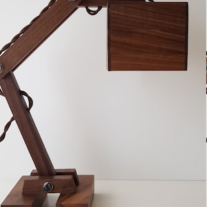 K&ouml;nnte beinhalten: Eine Schreibtischlampe aus Holz mit brauner Oberfl&auml;che. Die Lampe hat einen langen, beweglichen Arm und einen quadratischen Schirm. Die Lampe ist aus massivem Holz gefertigt und hat ein schlichtes, modernes Design.