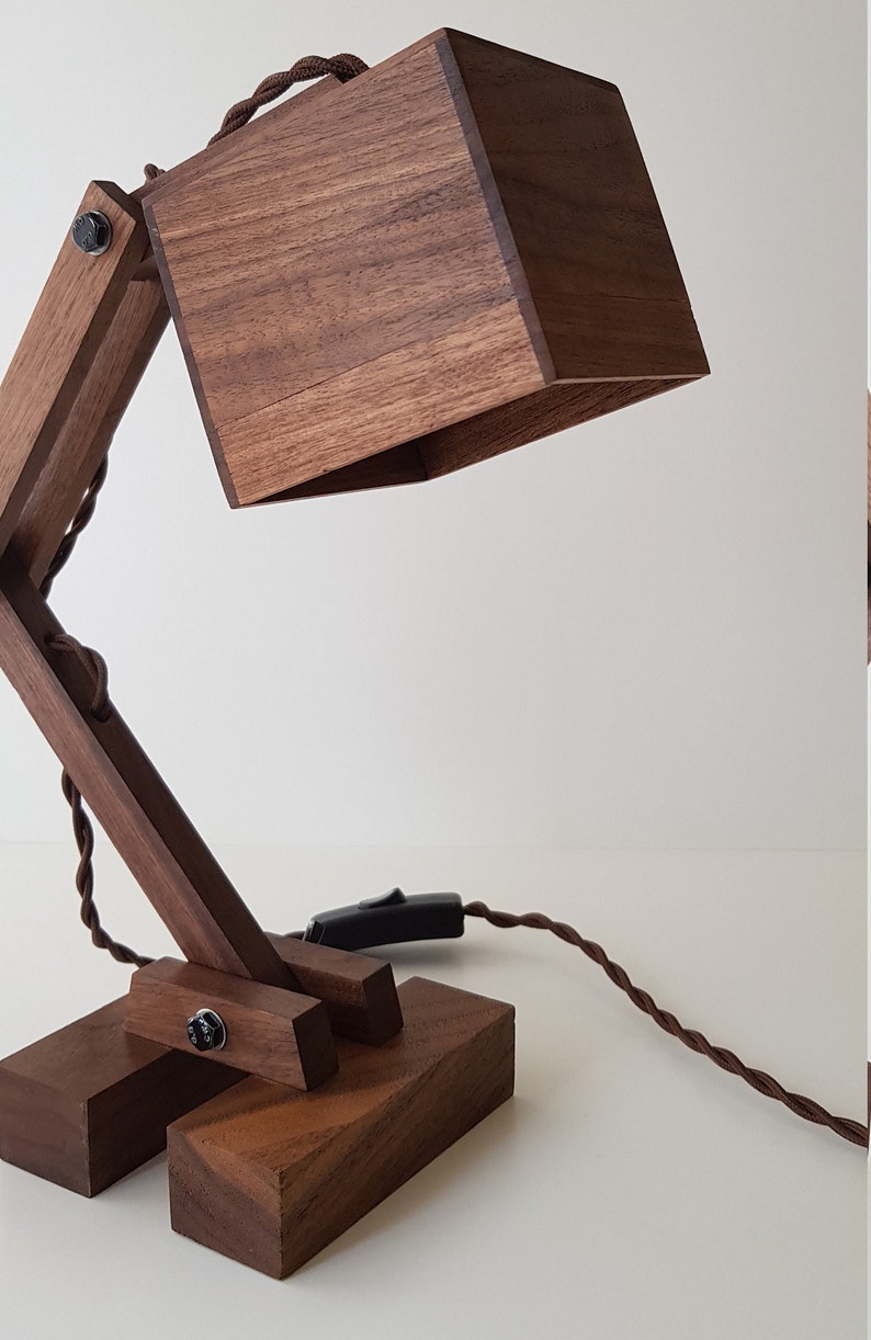 K&ouml;nnte beinhalten: Eine Schreibtischlampe aus Holz mit einem quadratischen Schirm und einem braunen Kabel. Die Lampe ist aus dunklem Holz gefertigt und hat ein schlichtes, modernes Design.