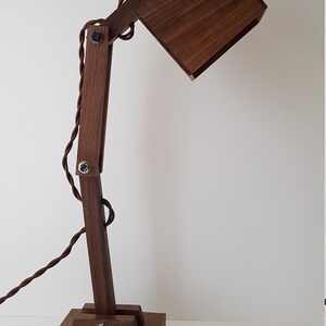 K&ouml;nnte beinhalten: Eine Schreibtischlampe aus Holz mit einem quadratischen Schirm und einem braunen Kabel. Die Lampe hat einen Gelenkarm und einen Sockel aus mehreren Holzst&uuml;cken.