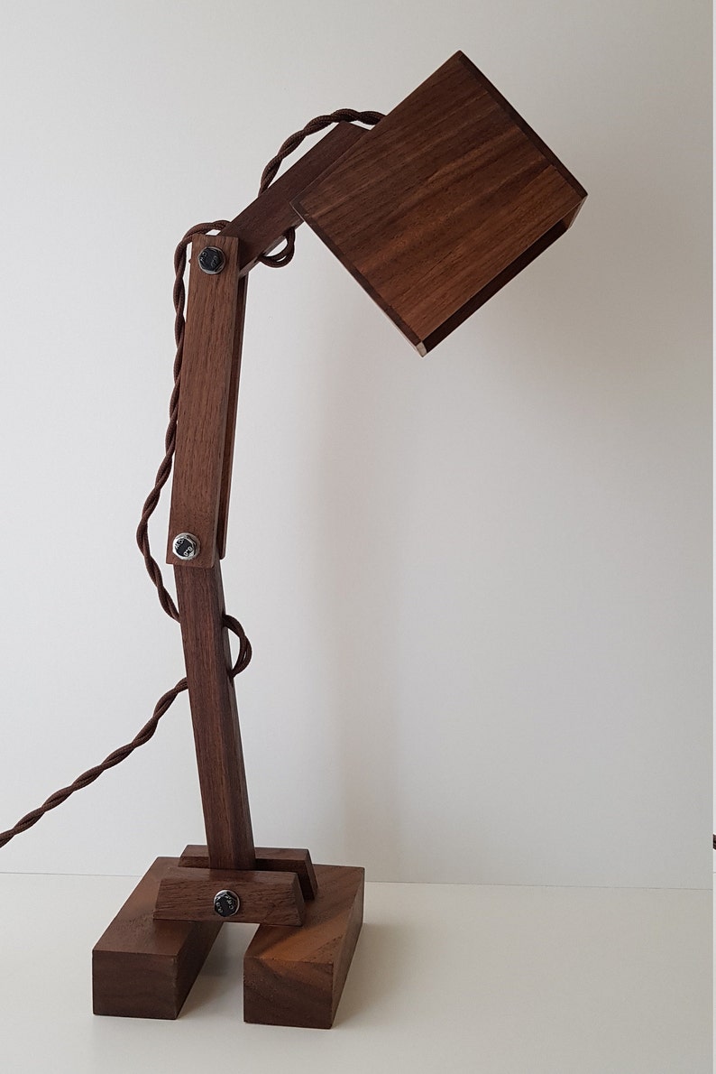 K&ouml;nnte beinhalten: Eine Schreibtischlampe aus Holz mit einem quadratischen Schirm und einem braunen Kabel. Die Lampe hat einen Gelenkarm und einen Sockel aus mehreren Holzst&uuml;cken.