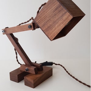 K&ouml;nnte beinhalten: Eine Schreibtischlampe aus Holz mit einem quadratischen Schirm und einem flexiblen Arm. Die Lampe ist aus dunklem Holz gefertigt und hat ein schlichtes, modernes Design.