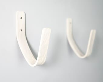 Puede incluir: Dos ganchos de pared de madera blanca en forma de U. Los ganchos están montados en una pared blanca.