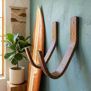 May include: Two wooden surfboard racks fixed to a teal wall. The racks are curved and have a dark brown finish. A surfboard with orange and white stripes leans against the wall. A potted plant and a woven wall hanging are also visible.