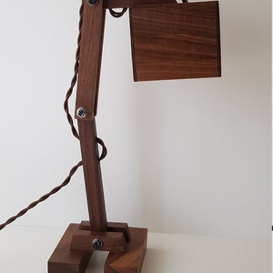 K&ouml;nnte beinhalten: Eine Schreibtischlampe aus Holz mit brauner Oberfl&auml;che. Die Lampe hat einen langen, beweglichen Arm und einen quadratischen Schirm. Die Lampe ist aus massivem Holz gefertigt und hat einen Vintage-Industriestyle.